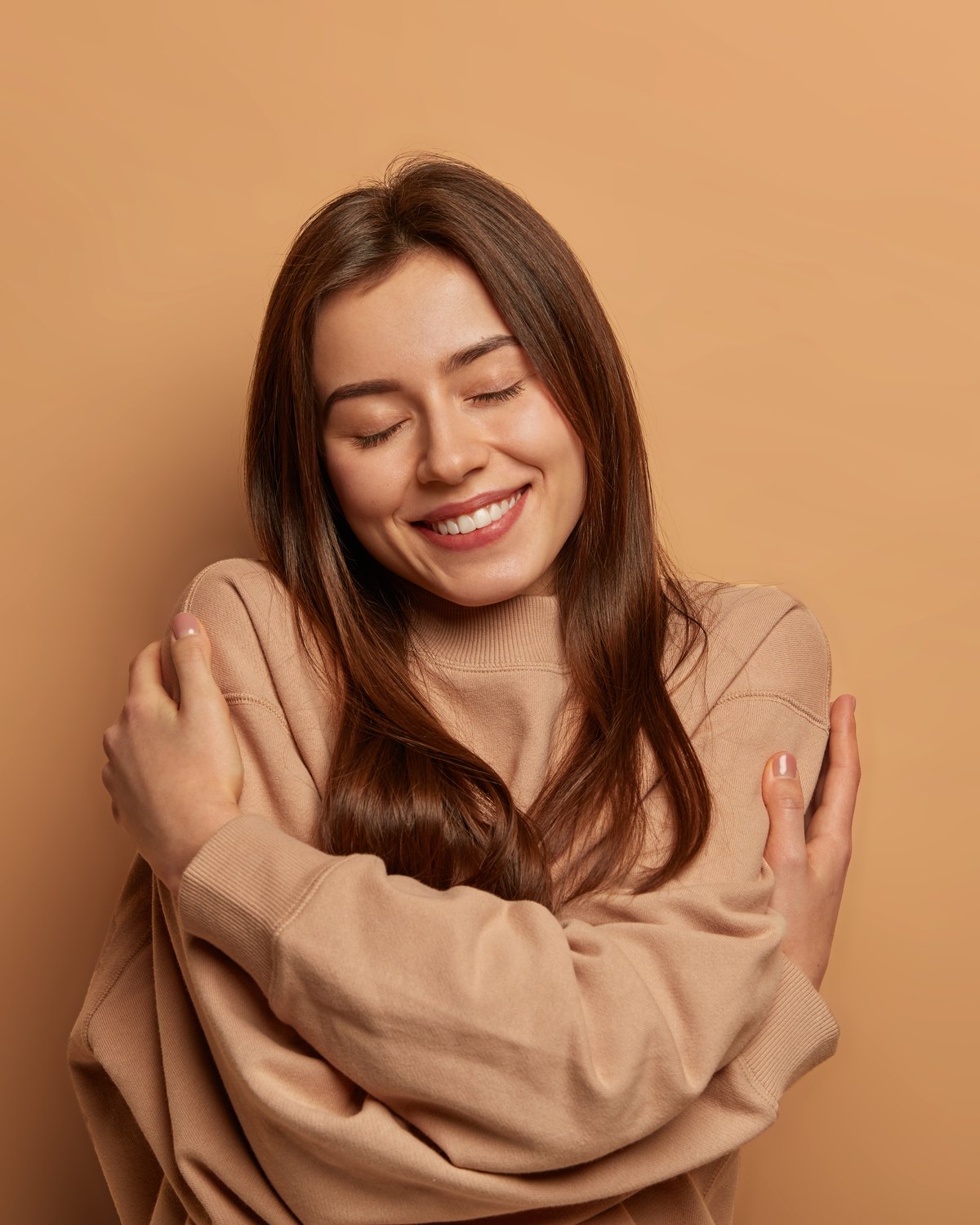 A woman smiling gently with her arms wrapped around herself, warm neutral background